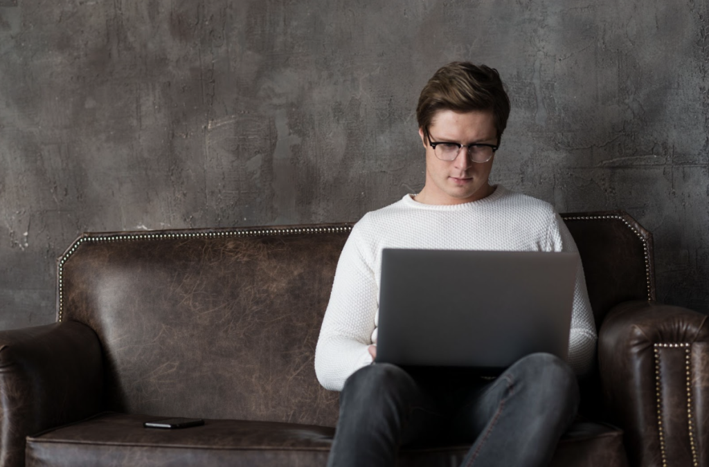 Une personne portant des lunettes et un pull blanc est assise sur un canapé en cuir marron, travaillant sur un ordinateur portable avec un smartphone posé à côté d'elle.