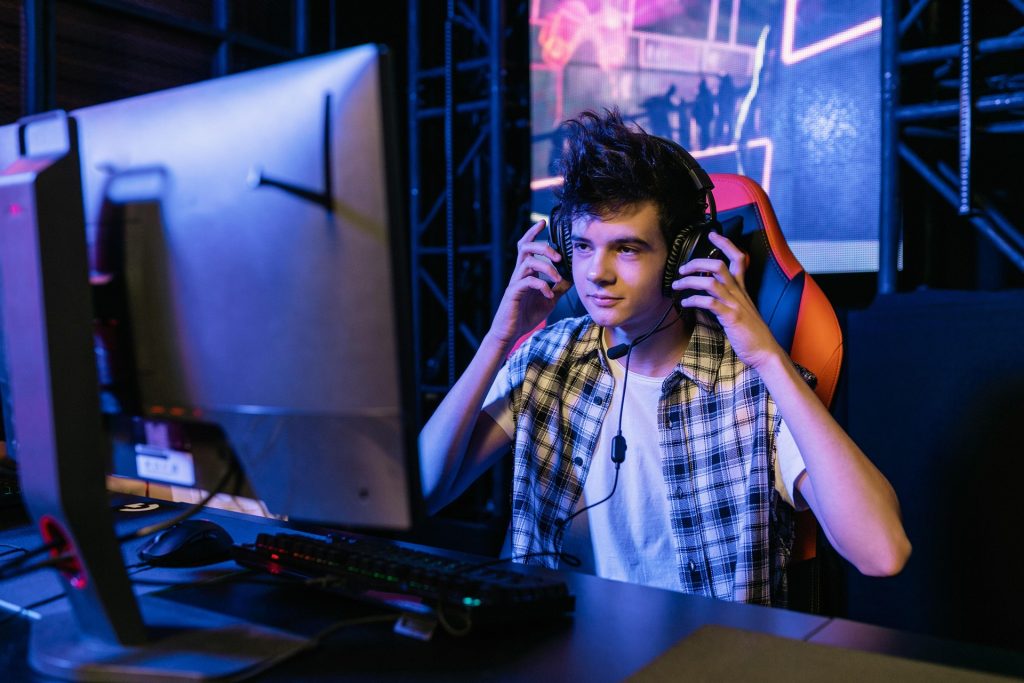 Un jeune portant des écouteurs est assis à un poste informatique, concentré sur l'écran, avec des équipements de jeu et des néons en arrière-plan.